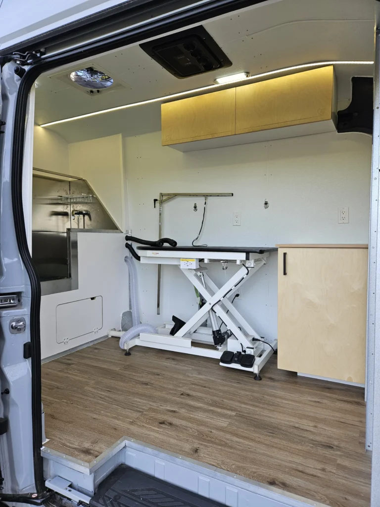 Interior of a grooming van with grooming table, wooden cabinets and wood-look flooring.