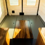 Interior of a small wooden room with three windows, featuring a central wooden table with a glossy surface and a dark edge, surrounded by a low black border, and two wooden countertops on either side.