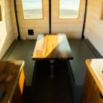 Interior of a small wooden camper with windows, a table in the center, and two wooden countertops.