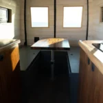 Inside a small wooden kitchen with three windows, a central table, wooden cabinets, and sunlight streaming in.
