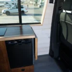 Interior of a camper van showing a small black refrigerator, a wooden countertop, near a window with a parking lot outside, and a black vehicle seat in the foreground.