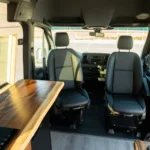 Interior of a camper van showing a wooden countertop, two front seats, and a dashboard with a steering wheel in the background.