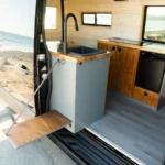 Interior of a tiny camper van with a small kitchen that includes a black sink, wooden countertops, a small refrigerator, and a window overlooking a river, parked on a beach.