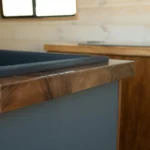 Close-up of a wooden countertop with a black sink inside a rustic kitchen or camper interior with wooden walls and a window.