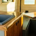 Inside a tiny home or camper with wood-paneled walls, a black sink, a wooden counter with a rounded edge, a small window, and a table with a striped wood pattern and a single white leg.